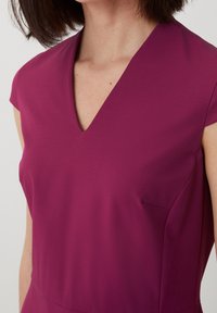 Fuchsia Kleid mit tiefem V-Ausschnitt, kurzen Puffärmeln und einer dezenten Seitennaht. Glattes Material mit einer minimalistisch geschnittenen, taillierten Silhouette.