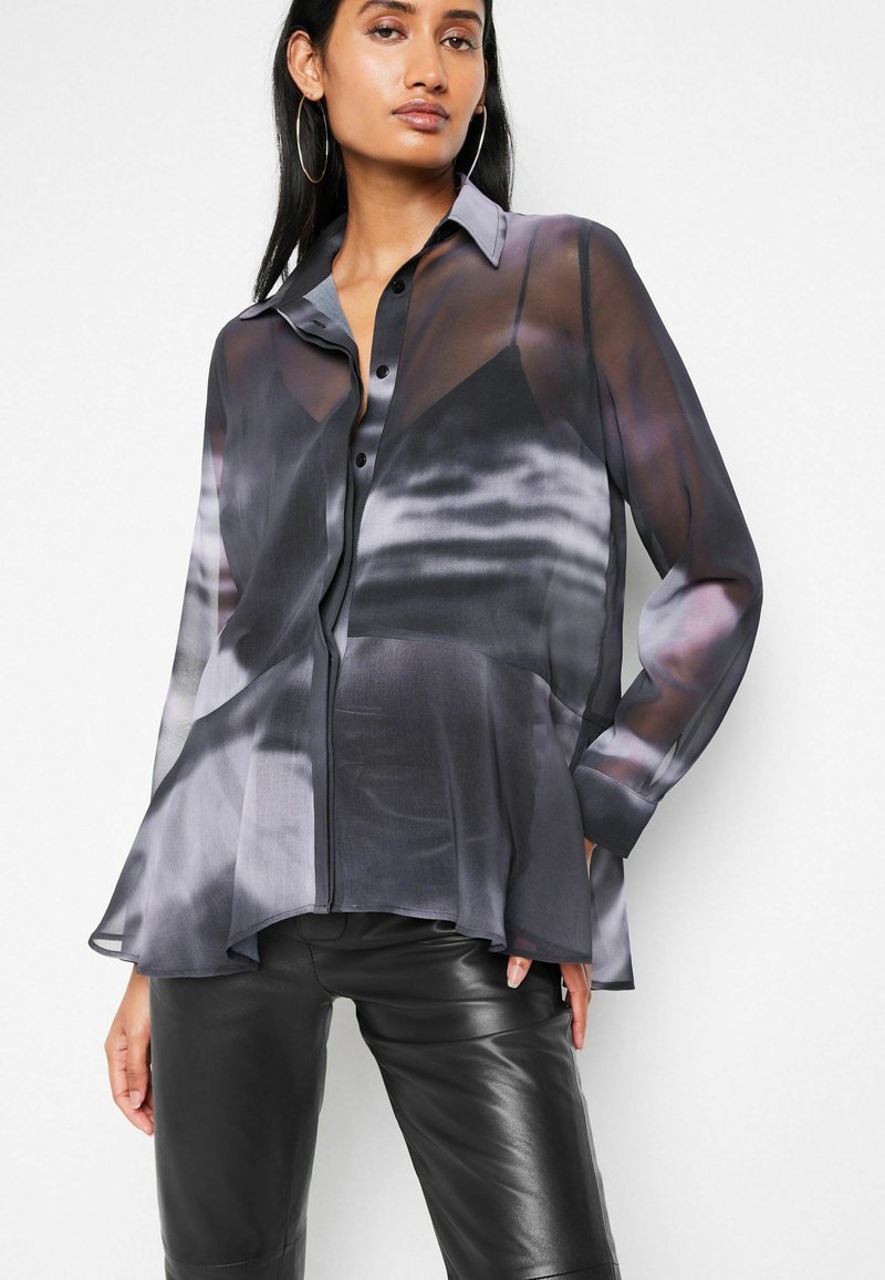 Sheer button-up shirt in gray with black gradient patterns; features a pointed collar and elongated hemline, paired with black leather pants.