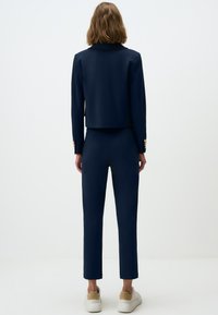 Navyblauwe cropped jack met een getailleerde pasvorm en een kraag, gecombineerd met bijpassende slim fit broek. Voorzien van gouden knopenaccenten en strakke witte sneakers.