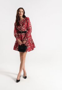 Czerwona sukienka w kwiaty z długimi rękawami i marszczoną talią, czarny pasek z złotym akcentem, w połączeniu z czarnymi szpilkami i małą torebką.