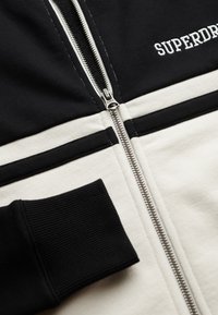 Sweat à capuche zippé noir et blanc avec poignets côtelés et logo bien visible. Tissu lisse avec motif en blocs de couleur horizontaux. Fermeture éclair.