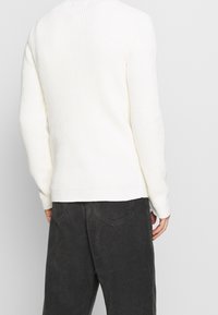 Weißer, gerippter Strickpullover mit langen Ärmeln, Rundhalsausschnitt und enganliegendem Saum, kombiniert mit dunklen Cordhosen.