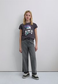 Camiseta gráfica gris con diseño de calavera y guitarra, combinada con jeans grises de pierna ancha con salpicaduras de pintura y zapatillas negras con estampado de leopardo.