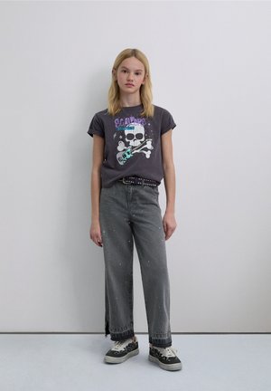 Camiseta gráfica gris con diseño de calavera y guitarra, combinada con jeans grises de pierna ancha con salpicaduras de pintura y zapatillas negras con estampado de leopardo.