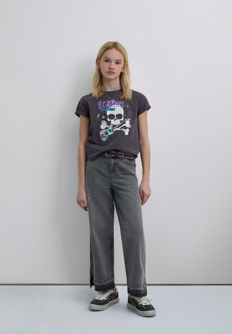 Camiseta gráfica gris con diseño de calavera y guitarra, combinada con jeans grises de pierna ancha con salpicaduras de pintura y zapatillas negras con estampado de leopardo.