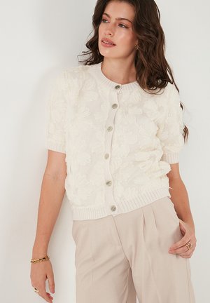 Cream cardigan with floral textured embroidery, short sleeves, ribbed neckline, and button closure. Smooth knit fabric and subtle sheen.
