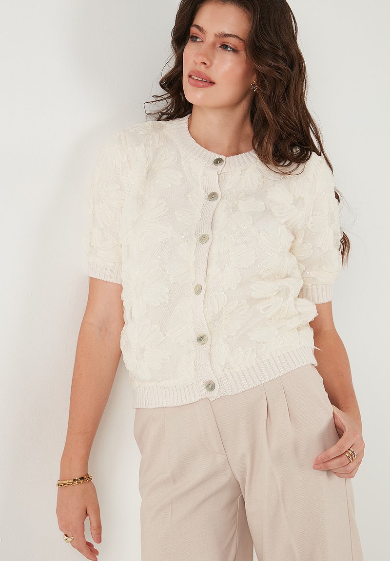 Cardigan crema con ricamo floreale a rilievo, maniche corte, scollo a costine e chiusura con bottoni. Tessuto a maglia morbida e leggero lucido.