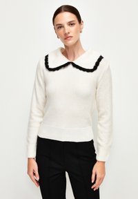 Pull en tricot blanc avec un col festonné noir et des poignets côtelés. La texture est douce et le design est ajusté. Associé à un pantalon noir.