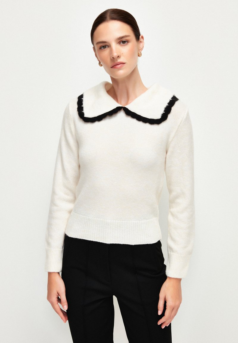 Pull en tricot blanc avec un col festonné noir et des poignets côtelés. La texture est douce et le design est ajusté. Associé à un pantalon noir.