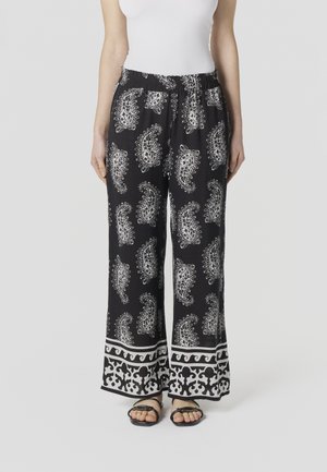 Mujer con pantalones anchos negros con estampado de cachemira blanco y un dobladillo estampado, combinados con sandalias negras de punta abierta y una blusa blanca.