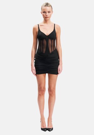 Femme aux cheveux blonds portant une robe body noire transparente avec une jupe froncée et des talons hauts noirs, debout devant un fond blanc uni.