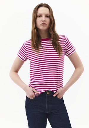 Młoda kobieta z długimi brązowymi włosami, ubrana w różowo-biały paskowany T-shirt i ciemnoniebieskie dżinsy, ręce w kieszeniach.
