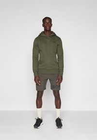 Felpa verde con cappuccio e pantaloni corti color khaki con un logo nero. Il modello indossa sneakers nere e calzini chiari. Abbigliamento sportivo semplice e informale.
