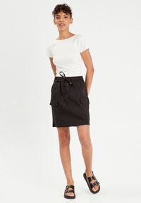 Jupe noire à cordon de serrage avec deux poches latérales, associée à un t-shirt blanc ajusté. Le tissu semble léger et confortable.