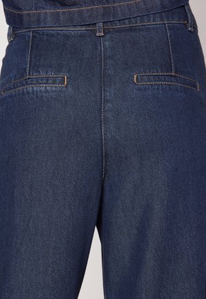 Un jean en denim bleu foncé présente une coupe classique, avec une texture lisse et des poches arrière accentuées par des coutures claires.