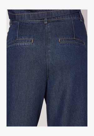 Jeans in denim blu scuro presentano un taglio classico, con una texture liscia e tasche posteriori accentuate da cuciture chiare.