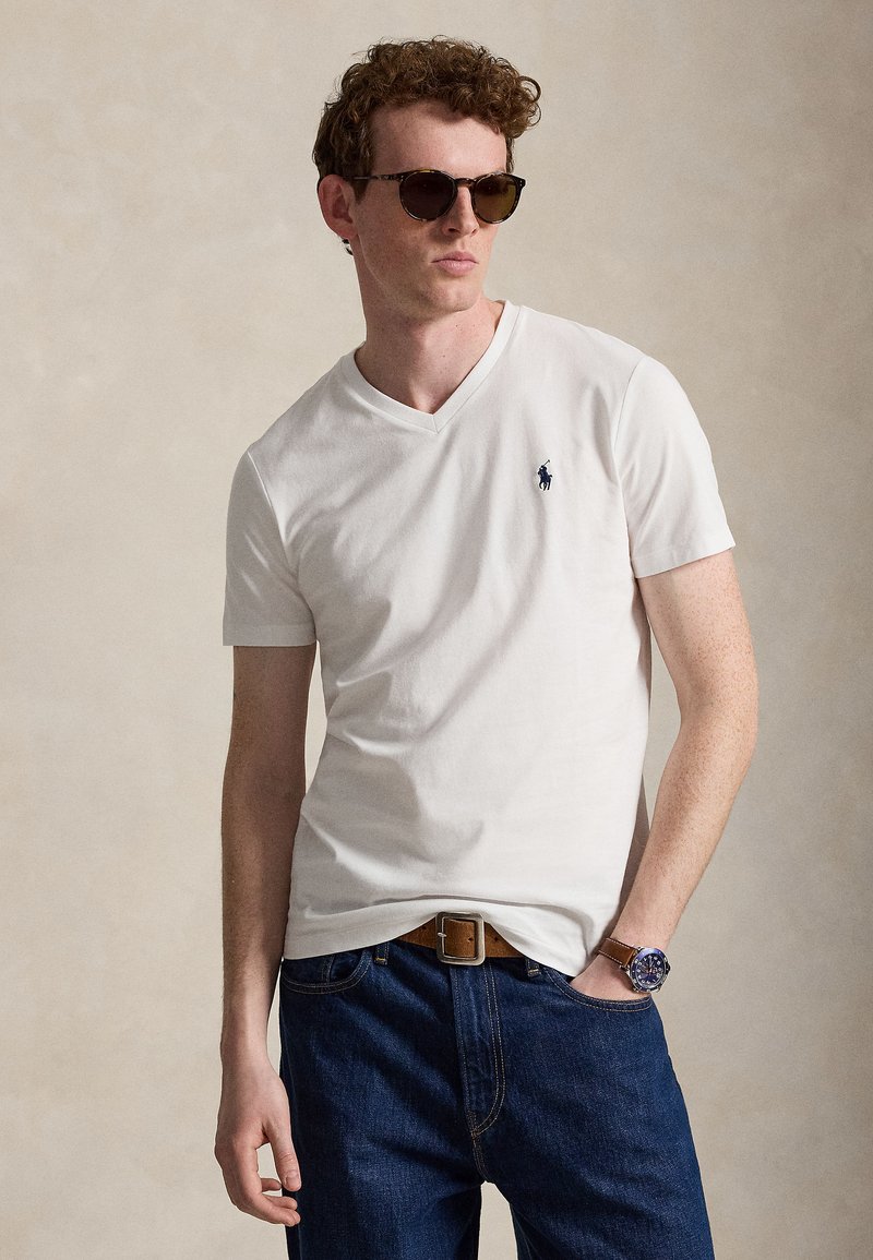 Mladý muž kučeravými vlasmi, ktorý nosí tmavé slnečné okuliare, biele tričko s výstrihom do V, modré džínsy, hnedý opasok a náramkové hodinky, stojí s rukou v vačku.