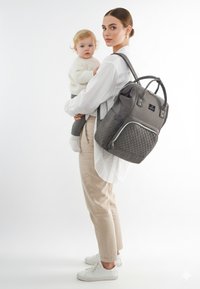 Grå quilted rygsæk med dobbelte top-håndtag, frontlomme med lynlås og justerbare stropper. En kvinde holder en baby i en cremefarvet sweater mod en lys baggrund.