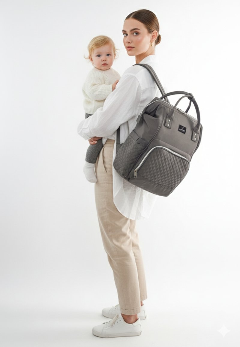 Grå quilted rygsæk med dobbelte top-håndtag, frontlomme med lynlås og justerbare stropper. En kvinde holder en baby i en cremefarvet sweater mod en lys baggrund.