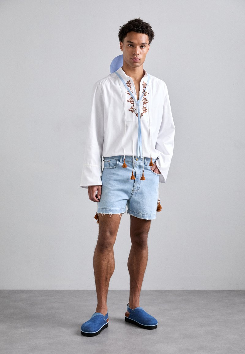 Camicia bianca a maniche lunghe con intricati ricami marroni e lacci blu, abbinata a pantaloni corti di jeans con dettagli a frange e scarpe slip-on blu.