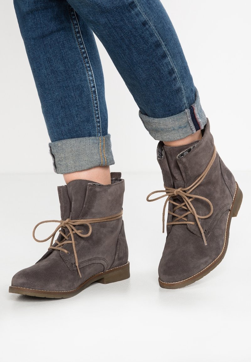 Botas de tobillo de ante gris con puntas redondeadas, diseño de cordones y suela de goma marrón baja, combinadas con unos vaqueros azules remangados.