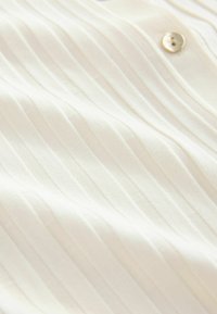 Camisa blanca con pliegues verticales, presenta una textura suave y un único botón transparente cerca del cuello. Acabado de tela limpio y suave.