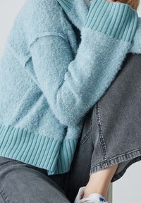 Personne portant un pull bleu clair texturé avec des poignets et un ourlet côtelés, associé à un jean en denim gris et des baskets blanches.