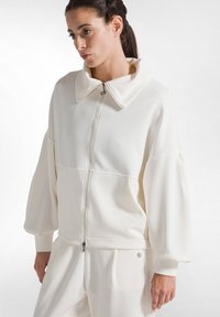 Chaqueta blanca con cremallera y gran cuello, mangas estructuradas y textura suave; combinada con pantalones a juego. Diseño y detalles minimalistas.