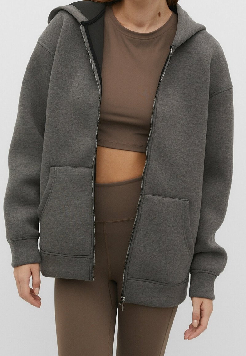 Sweat à capuche gris zippé en tissu doux, avec une capuche, deux poches avant et des poignets côtelés. Porté sur un crop top marron et un legging.