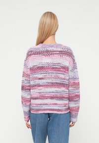 Person with long blonde hair wearing a multicolored striped knit sweater and blue jeans, standing facing a plain white background.