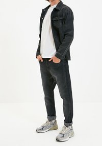 Giacca di denim nero sopra una camicia bianca, abbinata a jeans scuri e scarpe da ginnastica grigie. Look casual con maniche arrotolate e tasche.