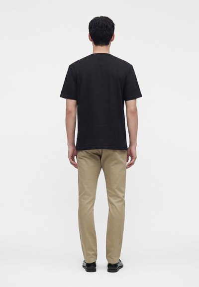 Homme de dos portant une chemise noire à manches courtes, un pantalon beige et des chaussures noires sur un fond clair uniforme.