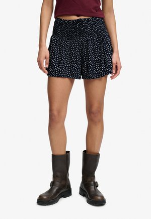 Kobieta nosząca czarne szorty z białym kwiatowym wzorem, w połączeniu z ciemnobrązowymi skórzanymi butami do połowy łydki, stojąca na białym tle.