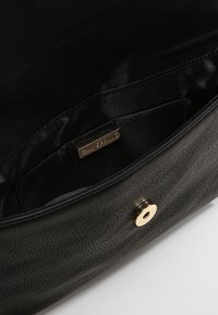 Pochette en cuir noir ouverte montrant l'intérieur en tissu noir avec une petite poche et une fermeture à bouton-pression magnétique doré.