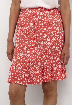 Czerwona spódnica na guziki z przodu, ozdobiona białymi wzorami kwiatowymi i abstrakcyjnymi, z falbanami na dole, noszona z białą bluzką wpuszczoną w spódnicę.