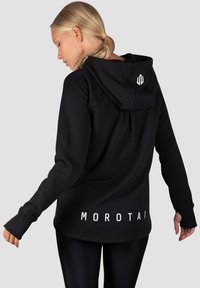 Čierna mikina s kapucňou vyrobená z mäkkého materiálu, s dlhými rukávmi, otvormi na palce a bielym logom na spodnej strane.
