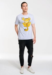 Man standing with hands behind back wearing a gray Garfield graphic t-shirt, black ripped jeans, and white sneakers on a white background.