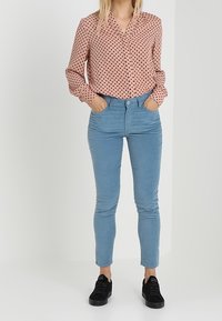 Mujer vestida con una blusa de lunares rosa de mangas largas, pantalones ajustados azul claro y zapatillas negras, con las manos en los bolsillos.