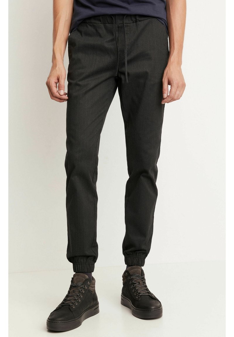 Homme portant un pantalon de jogging noir avec des poignets élastiques et des baskets montantes noires, debout sur un fond blanc.