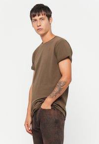 Brun bomull t-skjorte med korte ermer og rund hals, med en subtil logo på brystet og strukturert stoff, kombinert med mørke mønstrede jeans.