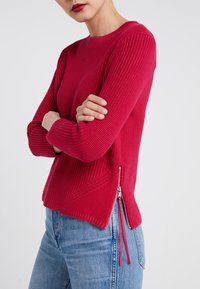 Sweter z dzianiny w jasnoróżowym kolorze o teksturowanej powierzchni, z okrągłym dekoltem, długimi rękawami i zamkami błyskawicznymi po bokach.