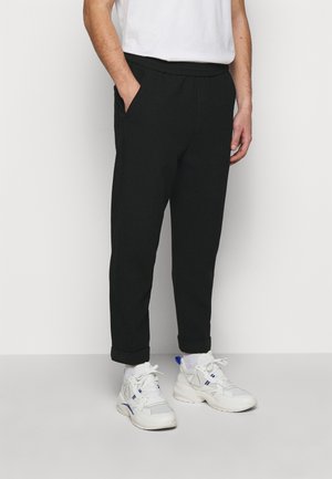 Man draagt zwarte taps toelopende broek met omgeslagen pijpen, witte sokken, witte chunky sneakers en een wit T-shirt met korte mouwen, staand tegen een effen achtergrond.