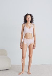 Jonge vrouw met vitiligo in lichtroze Calvin Klein ondergoed, staande op blote voeten op een vloerbedekking tegen een effen muur.