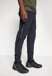 Marineblauwe sweatpants met ritszakken aan de zijkant en geribbelde manchetten, gecombineerd met zwarte sneakers voor atleten met textuurdetail en een reflecterende accent.