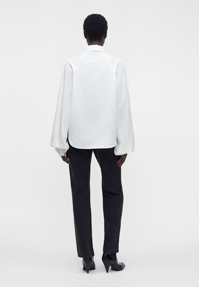 Personne debout de dos, portant un chemisier blanc à manches longues avec des poignets froncés, un pantalon noir et des chaussures à talons noires.