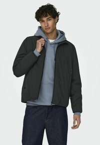 Black zippered jacket over a gray hoodie, featuring a relaxed fit, side pockets, and a textured fabric. Model stands with a casual pose.