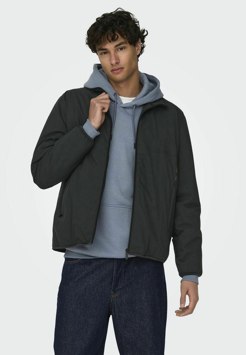 Black zippered jacket over a gray hoodie, featuring a relaxed fit, side pockets, and a textured fabric. Model stands with a casual pose.