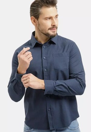 Mężczyzna poprawiający mankiet ciemnoniebieskiej koszuli na guziki, zwrócony w prawo, z krótkimi włosami i jasnym zarostem, noszący jasnoniebieskie dżinsy.