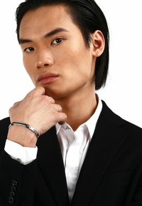 Bracciale d'argento indossato a un polso, caratterizzato da un design elegante e aerodinamico con accenti neri. Indossato da una persona in un abito nero e una camicia bianca.