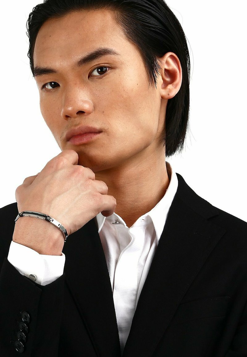 Bracciale d'argento indossato a un polso, caratterizzato da un design elegante e aerodinamico con accenti neri. Indossato da una persona in un abito nero e una camicia bianca.